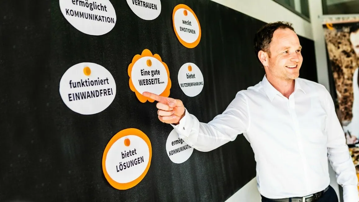 Foto von Sascha Krummeich während einer Präsentation. Er zeigt mit einem Finger auf einen Zettel mit der Aufschrift "funktioniert einwandfrei". Daneben hängen noch weitere Zettel.
